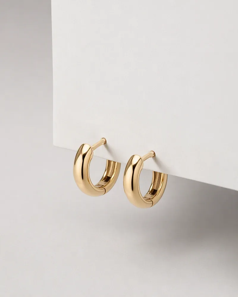 A pair of gold huggie hoops resting against a pale wall