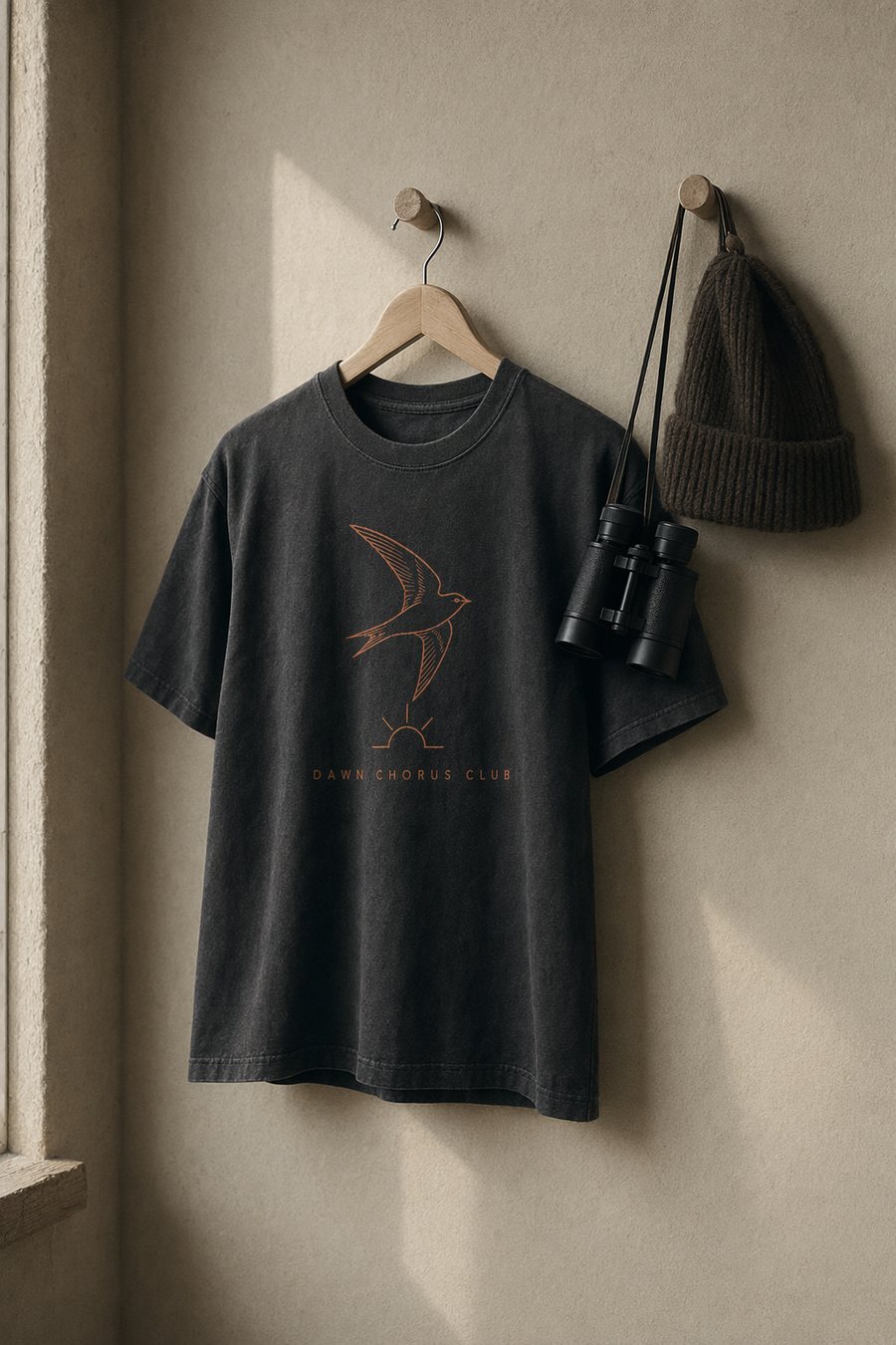 The 5AM Tee hanging on a wooden hanger beside binoculars
