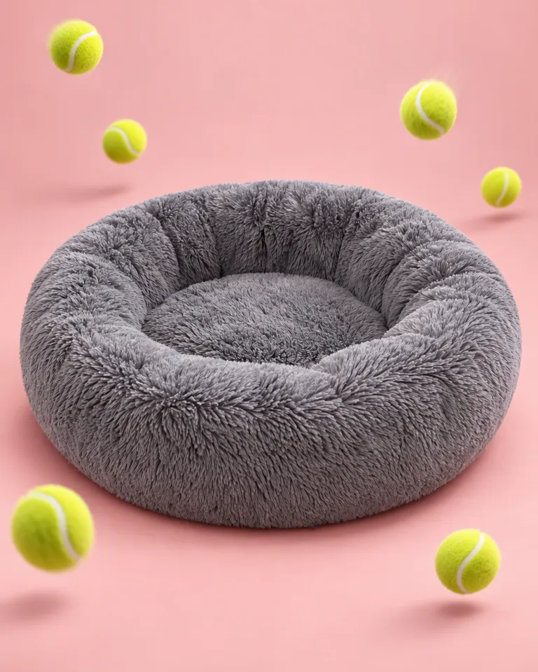 Cama de peluche gris para perro sobre un fondo rosa empolvado con pelotas de tenis congeladas en pleno bote